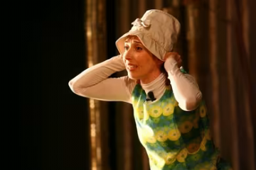scena con attrice vestita di verde che ha davanti a se una grande valigia di cuoio